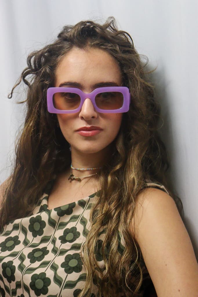 Femme portant une robe beige à imprimé floral vert, des lunettes de soleil violettes rectangulaires et un collier plaqué or. Look rétro chic signé Danie Hoo.