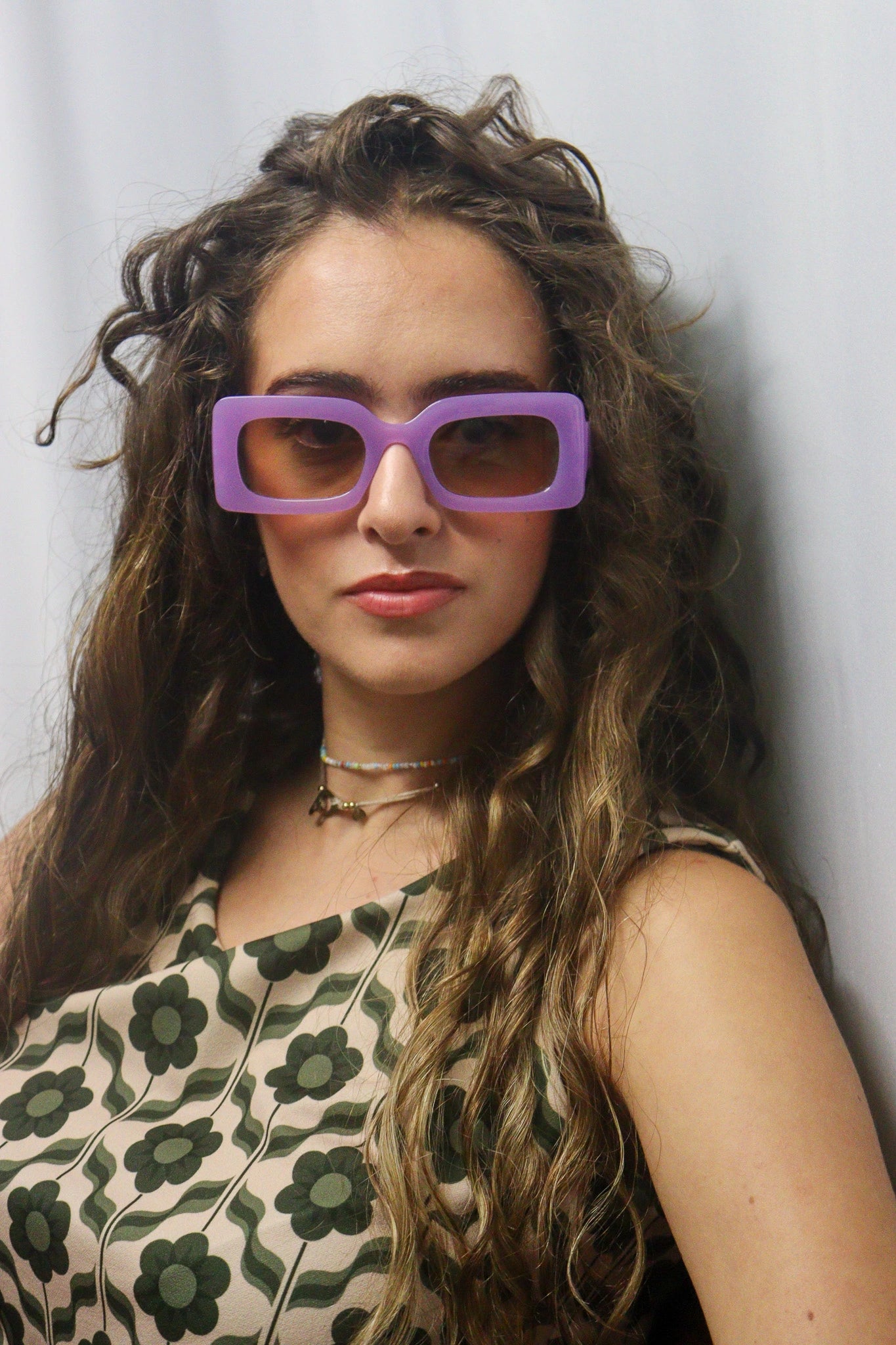 Jeune femme tendance portant des lunettes de soleil violettes rectangulaires oversize, un top sans manches à imprimé floral rétro vert sur fond beige, et des colliers fins. Look vintage chic, idéal pour le printemps-été.