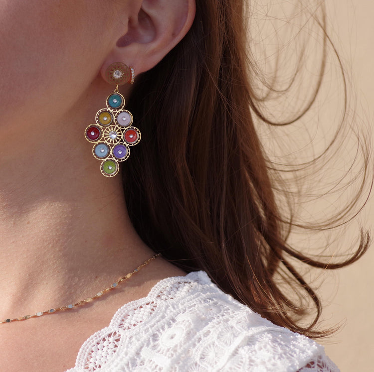 Femme portant une boucle d’oreille dorée ornée de pierres colorées et un collier fin assorti. Son haut en dentelle blanche apporte une touche bohème, tandis que le vent fait légèrement bouger ses cheveux.