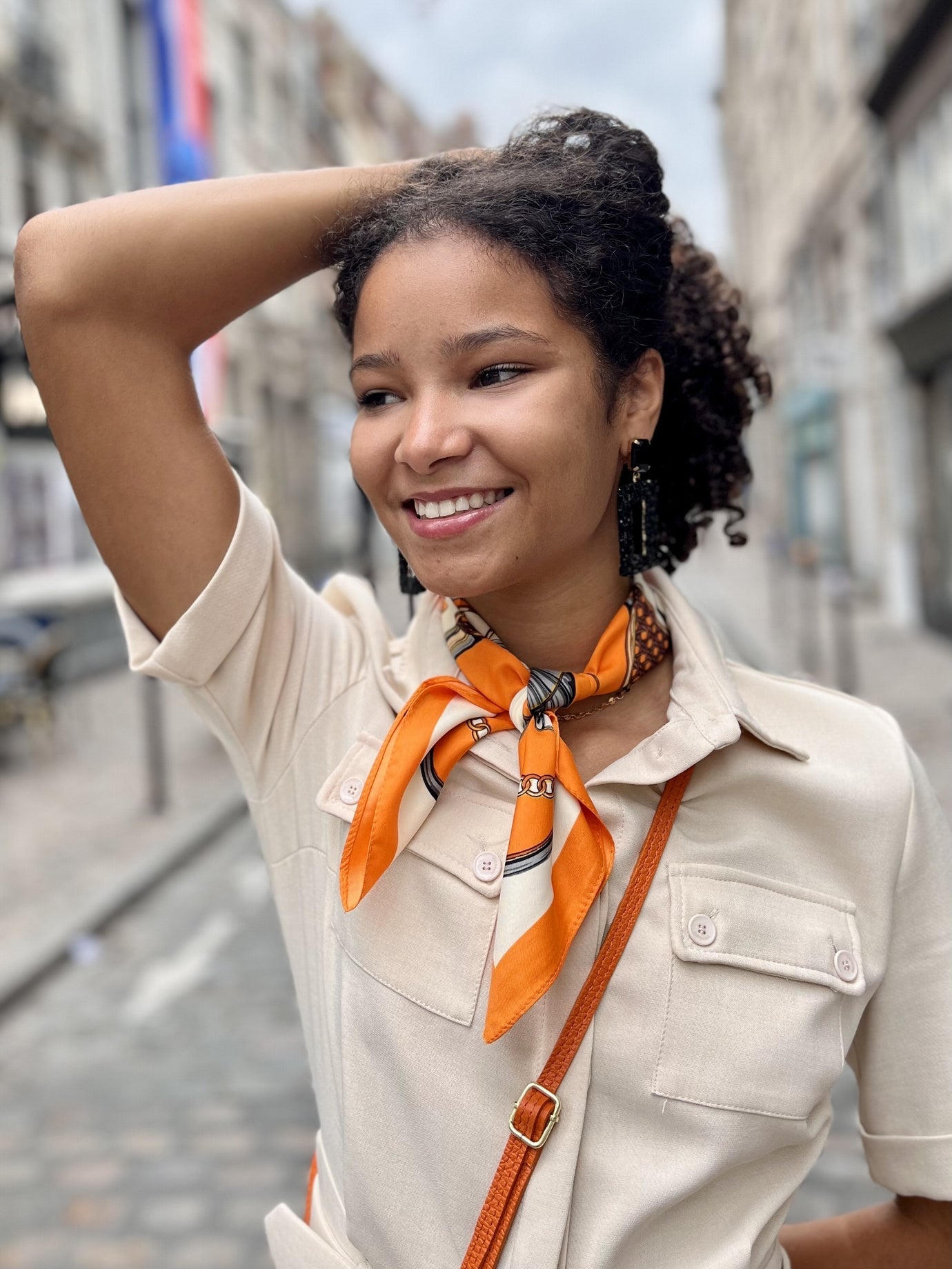 Jeune femme souriante portant un foulard orange noué autour du cou, une chemise beige et un sac en bandoulière assorti. Elle pose dans une rue pavée avec une main dans les cheveux, dégageant une allure élégante et décontractée.