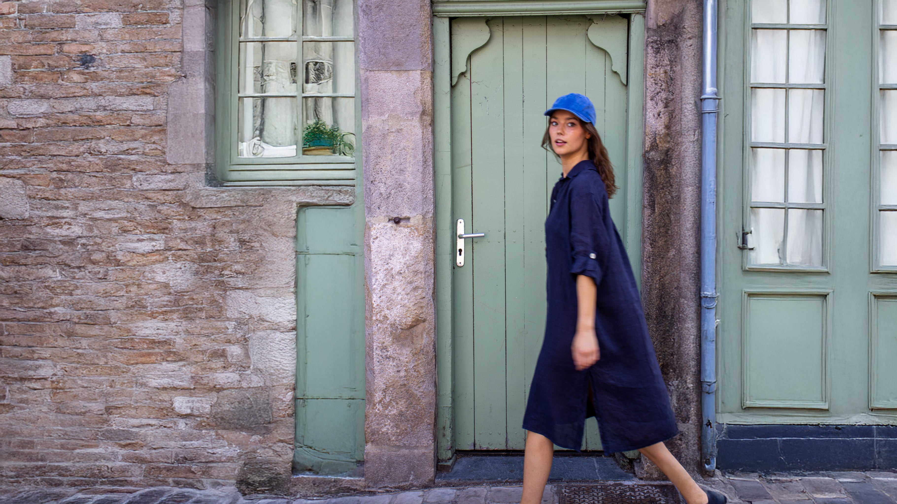 Instant capturé sur le vif dans une ruelle au charme ancien : une silhouette féminine traverse la scène avec élégance, vêtue d’une robe ample Danie Hoo. Ce modèle, dessiné sur la Côte d’Opale et fabriqué en Italie, incarne une mode à la fois libre, confortable et inspirée. Son bleu profond contraste avec la douceur des teintes pastel de la façade, pour une ambiance à la fois authentique et contemporaine.