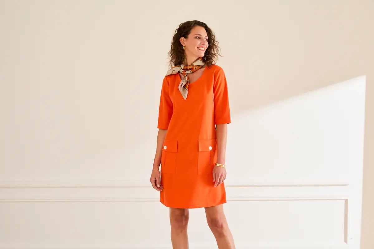 Femme souriante portant une robe orange élégante à manches mi-longues, posant dans un intérieur lumineux — collection été 2025.